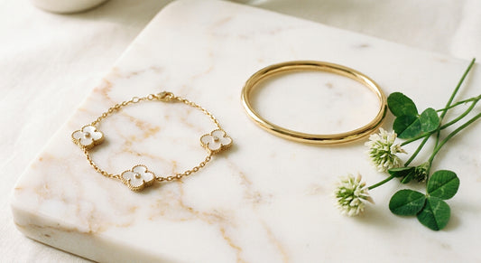 Two elegant bracelets — a clover-motif bracelet and a minimalist bangle — displayed side by side on a marble surface with soft natural lighting.