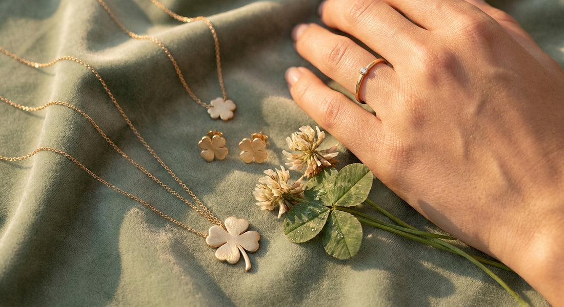 A woman's hand adorned with delicate gold four-leaf clover jewelry resting on sage green velvet alongside a real clover sprig in warm golden light.