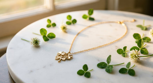 A gold-plated multi clover necklace displayed on white marble with soft natural lighting and scattered green clover leaves nearby.