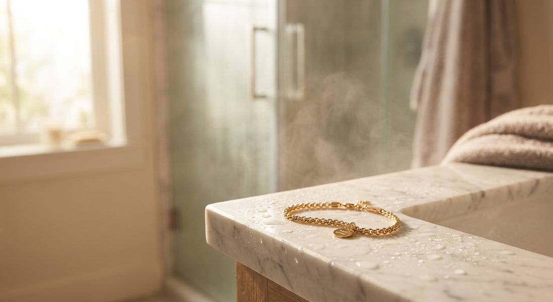 A gold chain bracelet resting on a marble bathroom surface with water droplets nearby and a softly blurred steamy shower in the background.