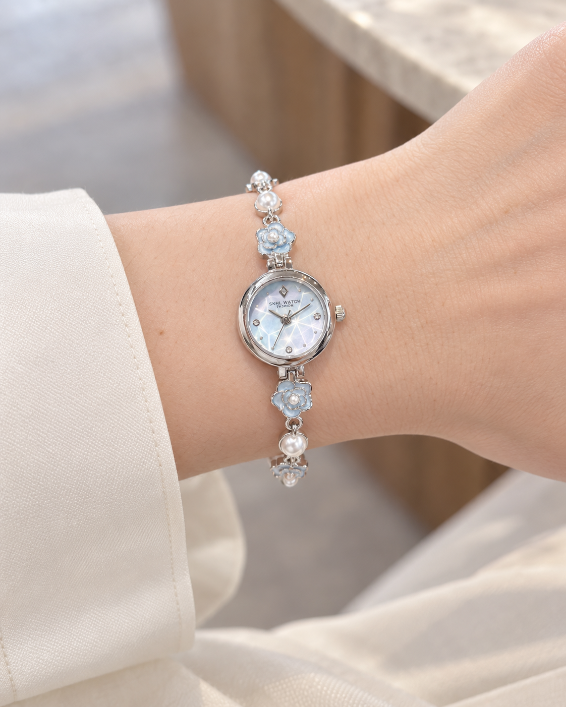 Blue dial silver-tone SKHL camellia bracelet quartz watch worn on an adult wrist with a cream blouse