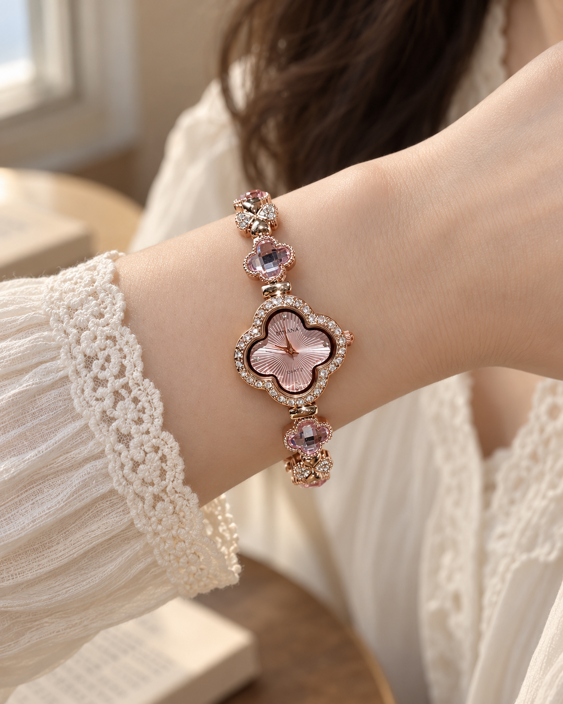 Pink AIMANA clover bracelet watch worn on an adult wrist with a cream lace sleeve