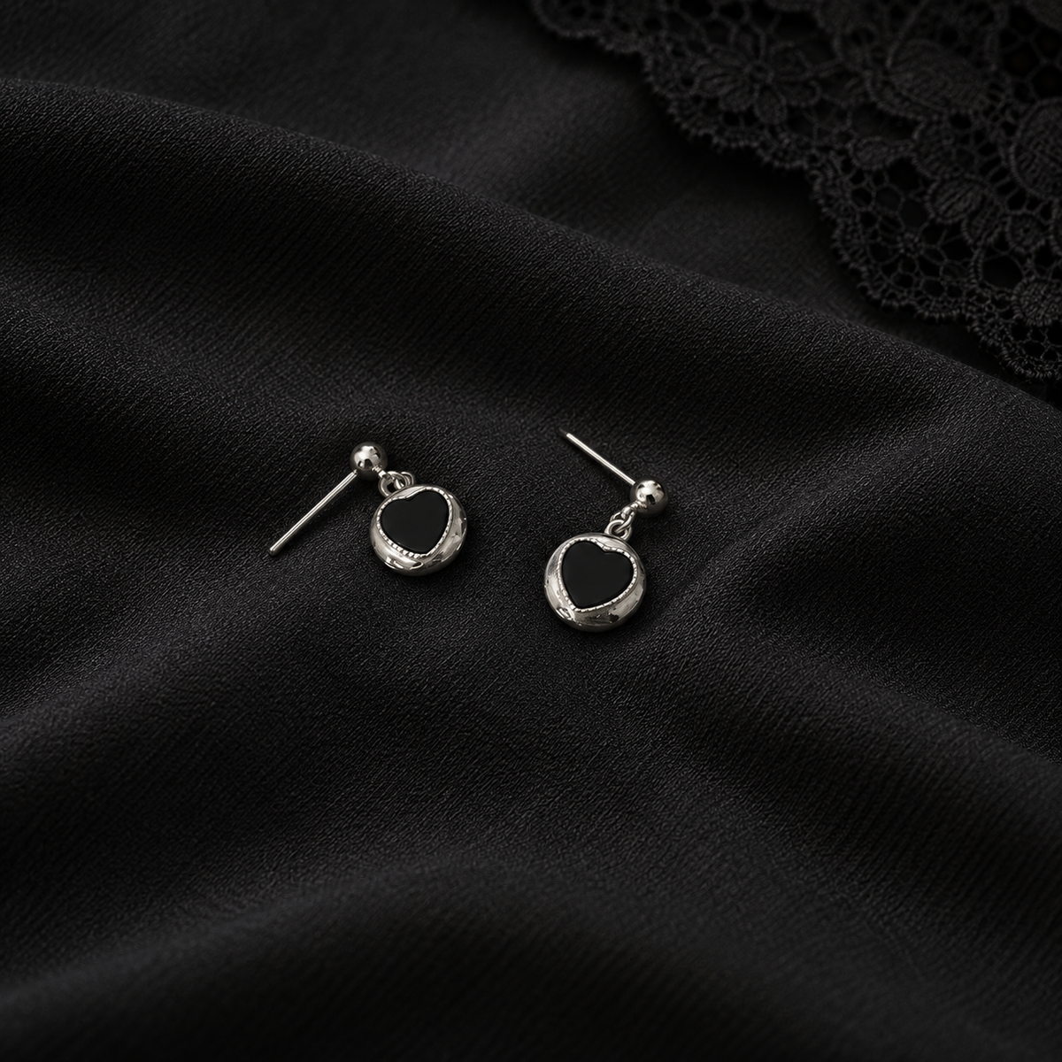 Silver-tone black heart drop earrings on textured black fabric with a lace edge